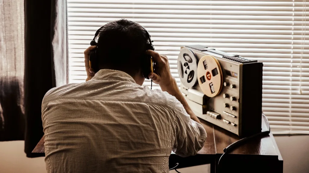 A man in headphones at a desk, listening to a reel-to-reel tape recorder beside backlit window blinds — a surveillance-style illustration.